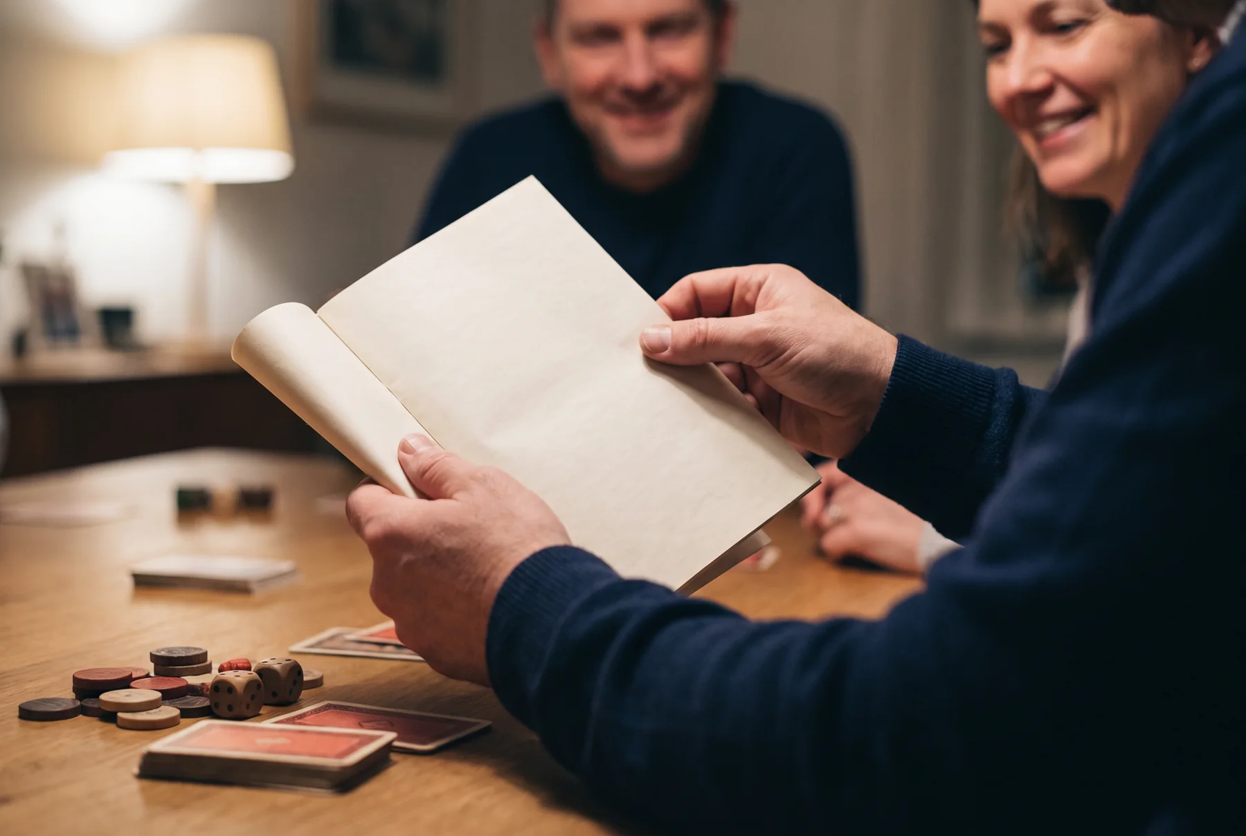 Host opening a blank rulebook beside tabletop game pieces